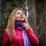 A woman in a red coat and purple scarf poses thoughtfully in a forest.